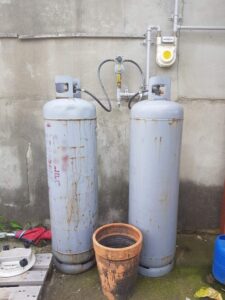 Two vertical propane tanks stand against a concrete wall with visible rust and wear, indicating outdoor use and weathering. An aged terracotta pot rests in the foreground, adding a sense of everyday outdoor utility to the scene.